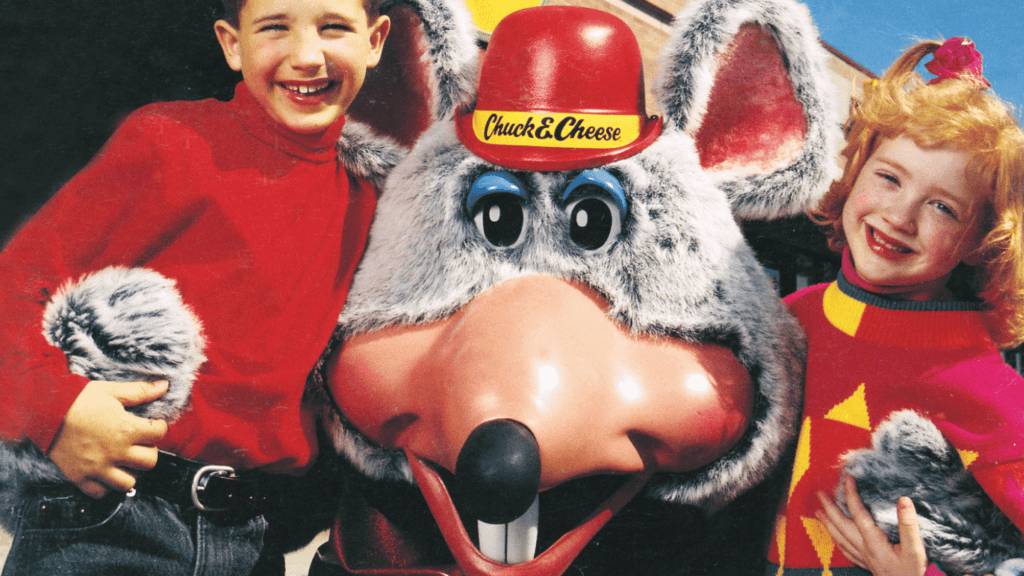 Two kids in colorful sweaters stand by Chuck E. Cheese mascot with red hat at family fun center, perfect for birthday party for kids.