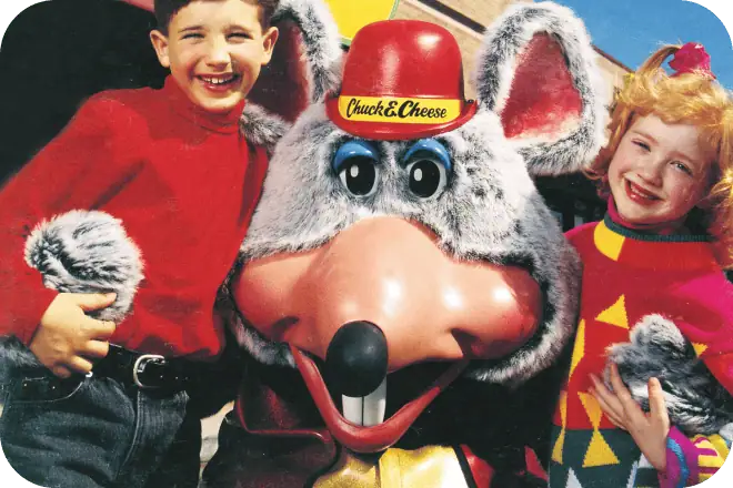 Smiling kids at a Chuck E. Cheese birthday party pose with Chuck E. Mouse, surrounded by arcade games for kids and pizza.