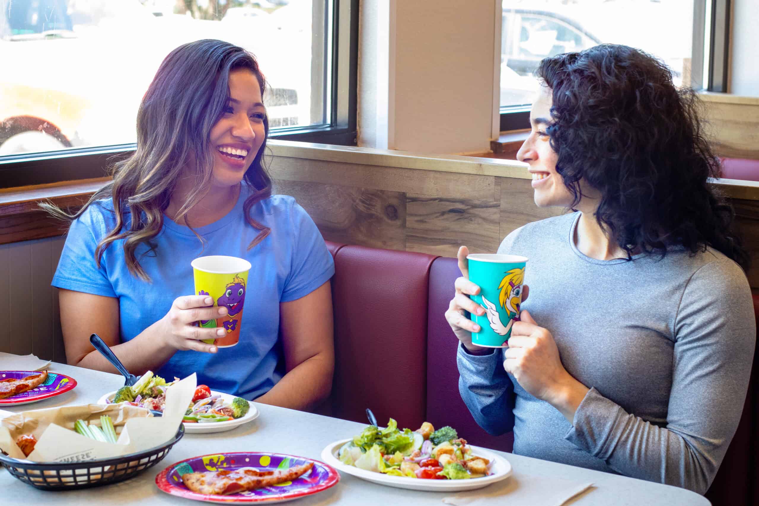 Two women enjoy salad and pizza at a Chuck E. Cheese booth, holding cups, with daylight and arcade games for kids in the background.