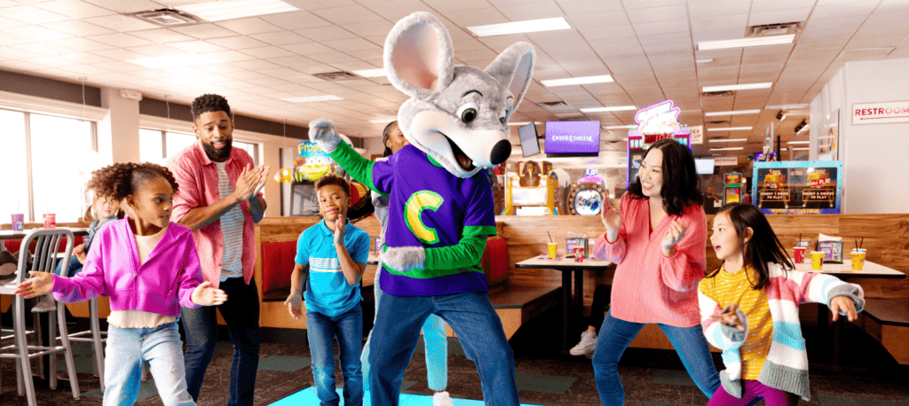 Kids dance with Chuck E. in purple shirt at Chuck E. Cheese; arcade games, pizza, tables, and Fun Pass visible at birthday party.