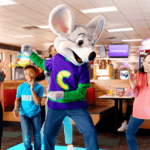 Kids dance with Chuck E. in purple shirt at Chuck E. Cheese; arcade games, pizza, tables, and Fun Pass visible at birthday party.