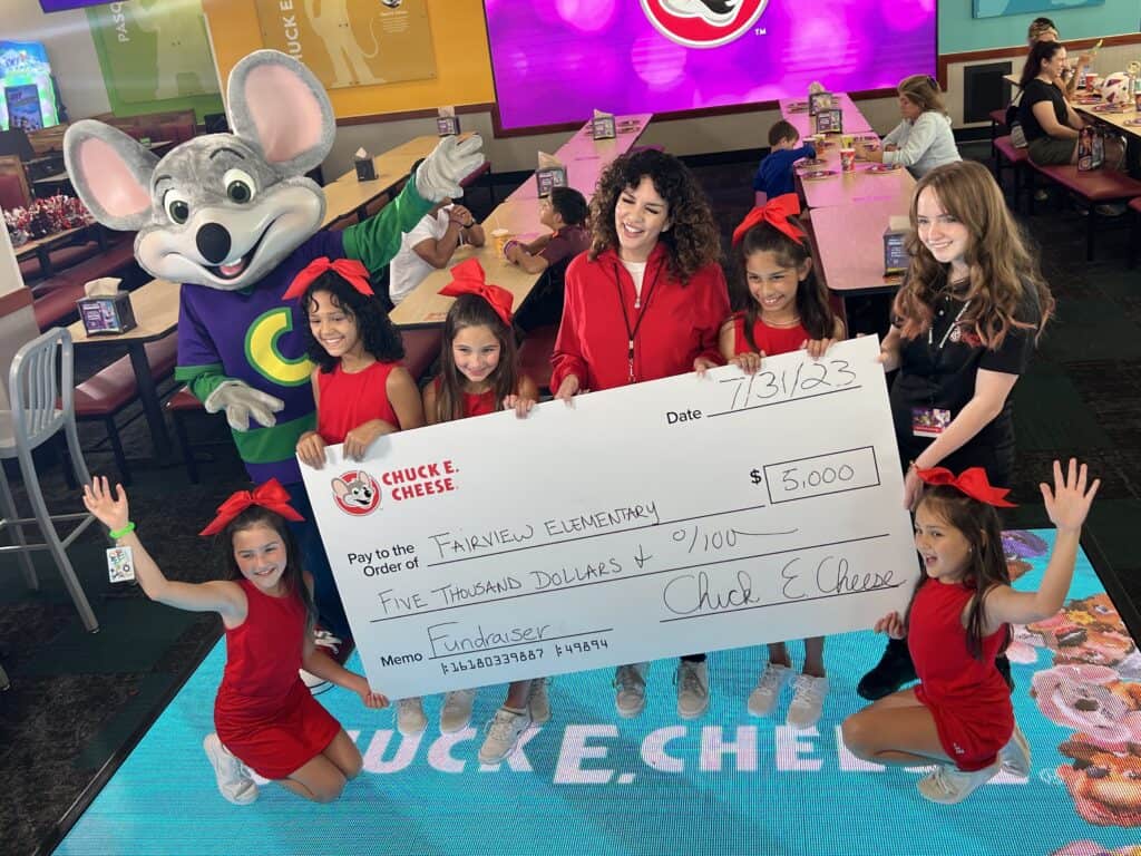 Five kids in red and three women hold a $5,000 check with Chuck E. Cheese at a family fun center with arcade games for kids.