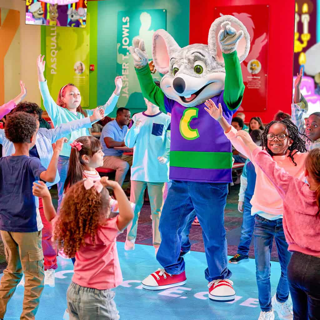 Children dance at a Chuck E. Cheese birthday party with the mascot; arcade games, pizza, and Fun Pass signs fill the family fun center.