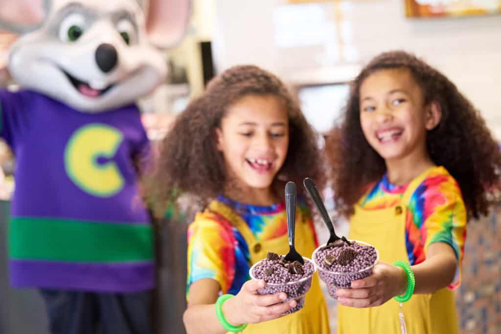 Two girls in yellow overalls eat ice cream at a Chuck E. Cheese birthday party, mascot and arcade games for kids in background.