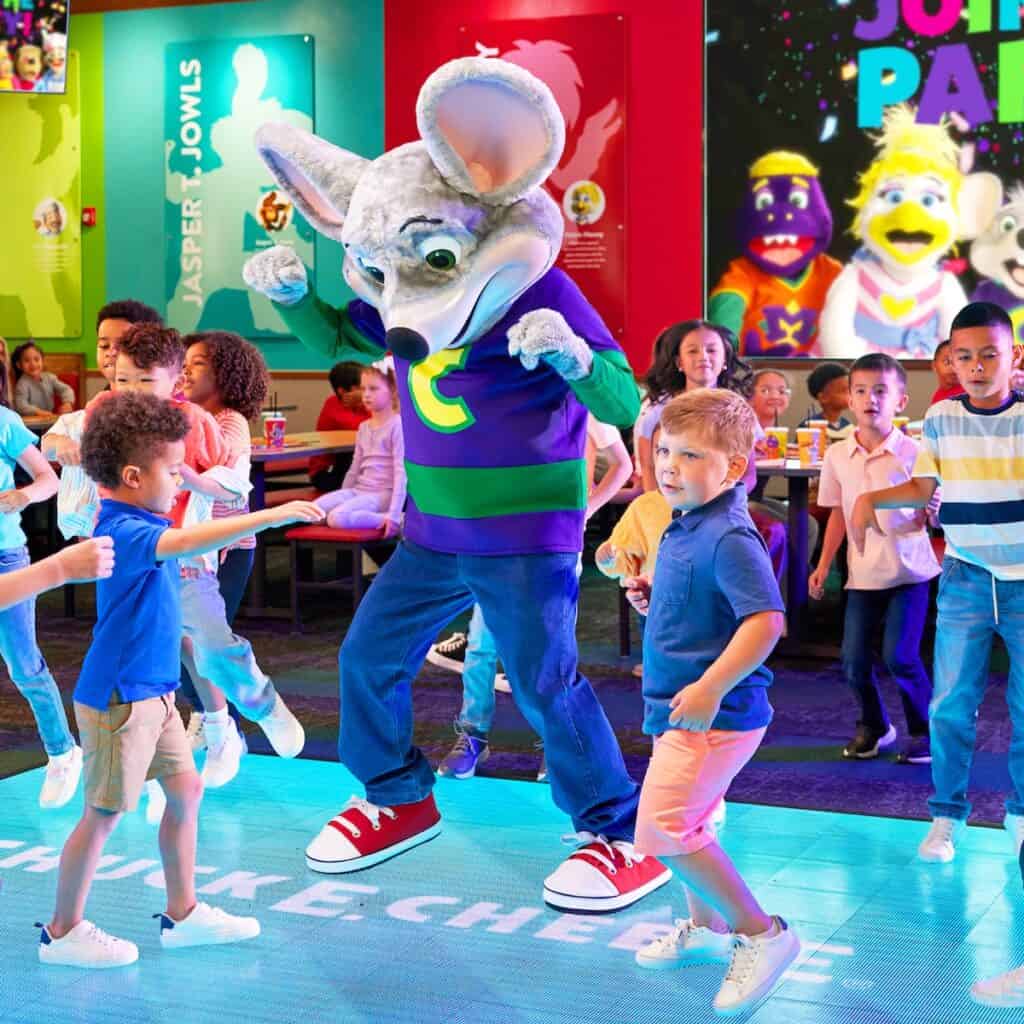 Chuck E. Cheese mascot dances with kids during a birthday party in play area, surrounded by arcade games, pizza, and Fun Pass cards.