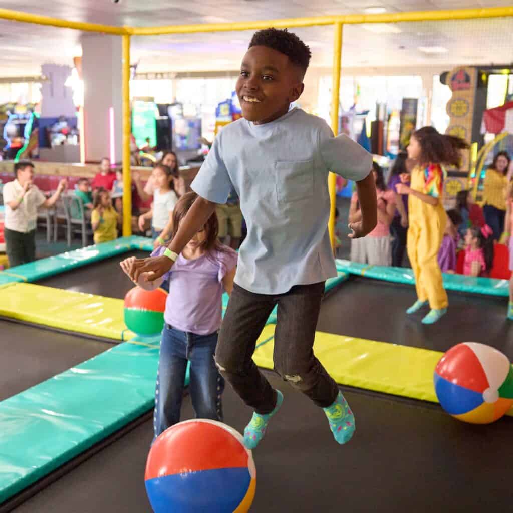 A boy in a light blue shirt jumps on a trampoline with beach balls at Chuck E. Cheese, near arcade games during a kids birthday party.