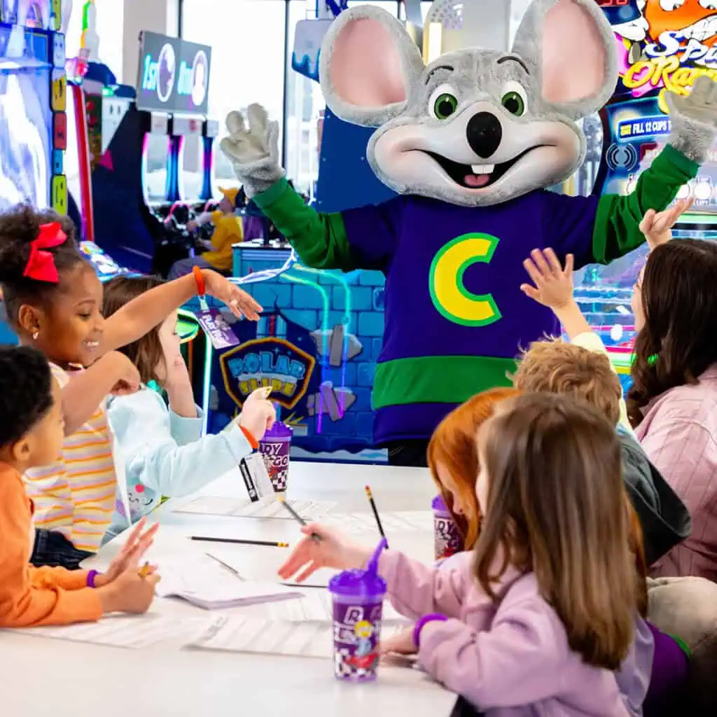 Kids at a Chuck E. Cheese birthday party sit around a table with pizza, raising hands as Chuck E. stands behind; arcade games visible.