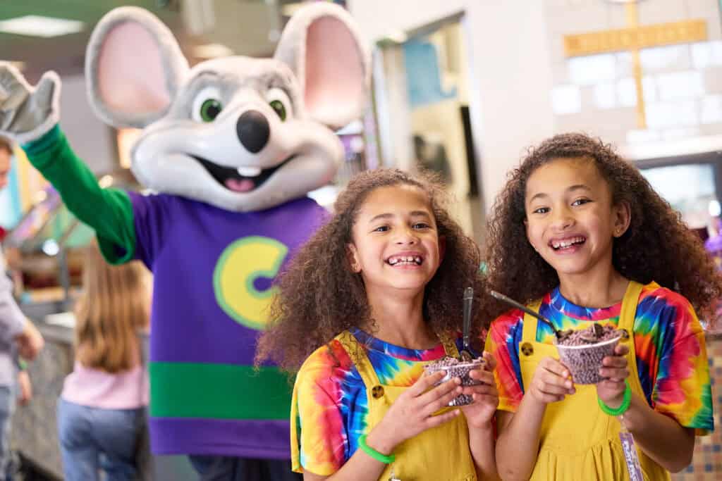 Two girls with ice cream stand by Chuck E. Cheese mascot in a vibrant family fun center with arcade games and birthday party decor.