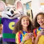 Two girls with ice cream stand by Chuck E. Cheese mascot in a vibrant family fun center with arcade games and birthday party decor.