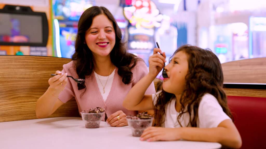 Woman and young girl enjoy chocolate ice cream with their Fun Pass at Chuck E. Cheese, surrounded by arcade games for kids.