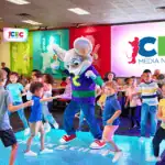Kids dance on a lit-up floor with Chuck E. Cheese at a birthday party, families watching in the family fun center with arcade games.