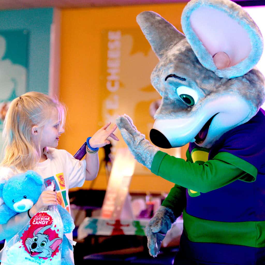 Girl with blue teddy bear and cotton candy high-fives Chuck E. Cheese mascot at birthday party in family fun center with arcade games.