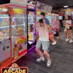 Teen boy at Chuck E. Cheese family fun center, frustrated at claw machine; kids play arcade games nearby, Chuck’s Arcade logo visible.