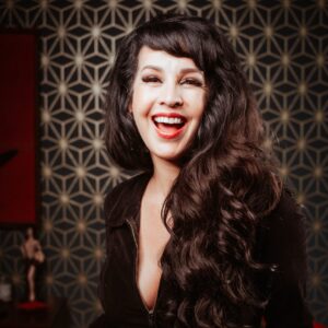 A woman with long, wavy dark hair smiles in front of a geometric wall at Chuck E. Cheese, known for kids’ birthday parties and arcade games.