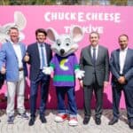 Six men in business attire with Chuck E. Cheese mascot at Türkiye family fun center backdrop, thumbs up for kids birthday party and games.