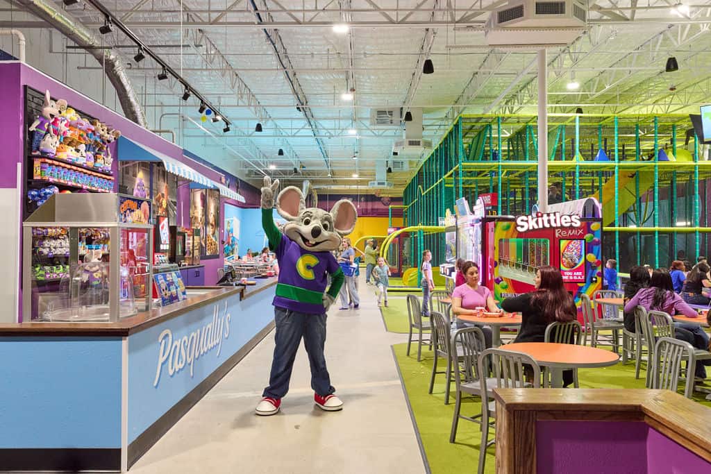 Chuck E. Cheese mascot greets kids at birthday party; arcade games, pizza, families, and a play structure fill the family fun center.