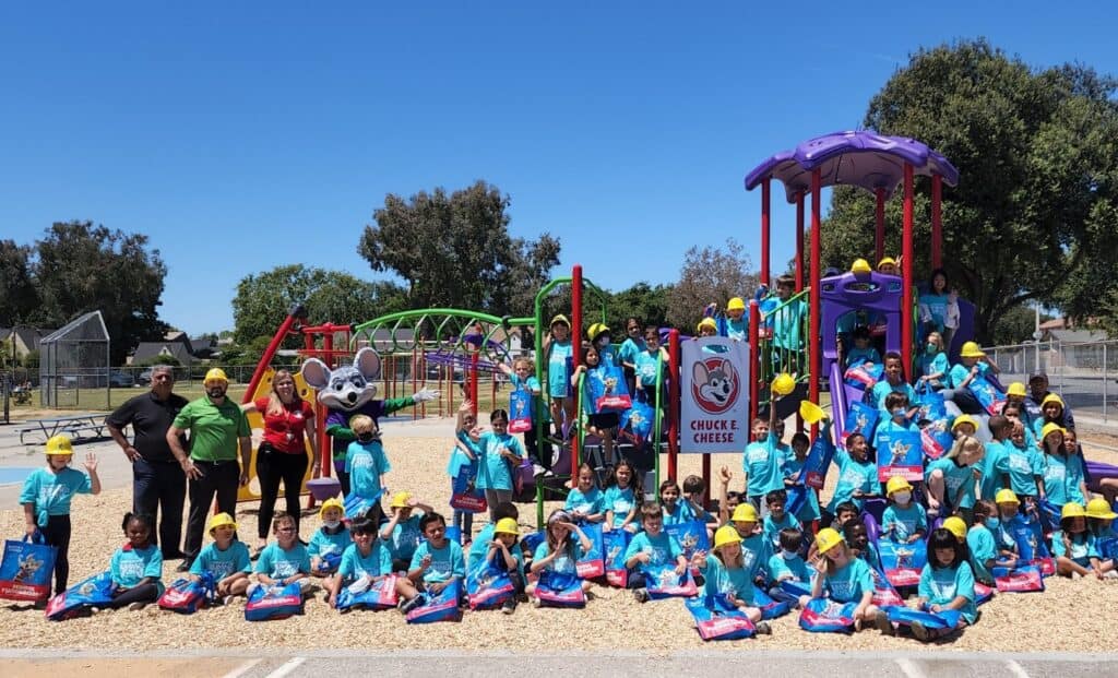 Kids in blue shirts and yellow hats with Chuck E. Cheese mascot at a birthday party, holding prize bags by arcade games and playground.