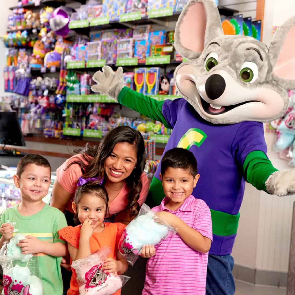 A woman and three kids with cotton candy stand by Chuck E. Cheese mascot in the arcade, toy shelves and games in the background.