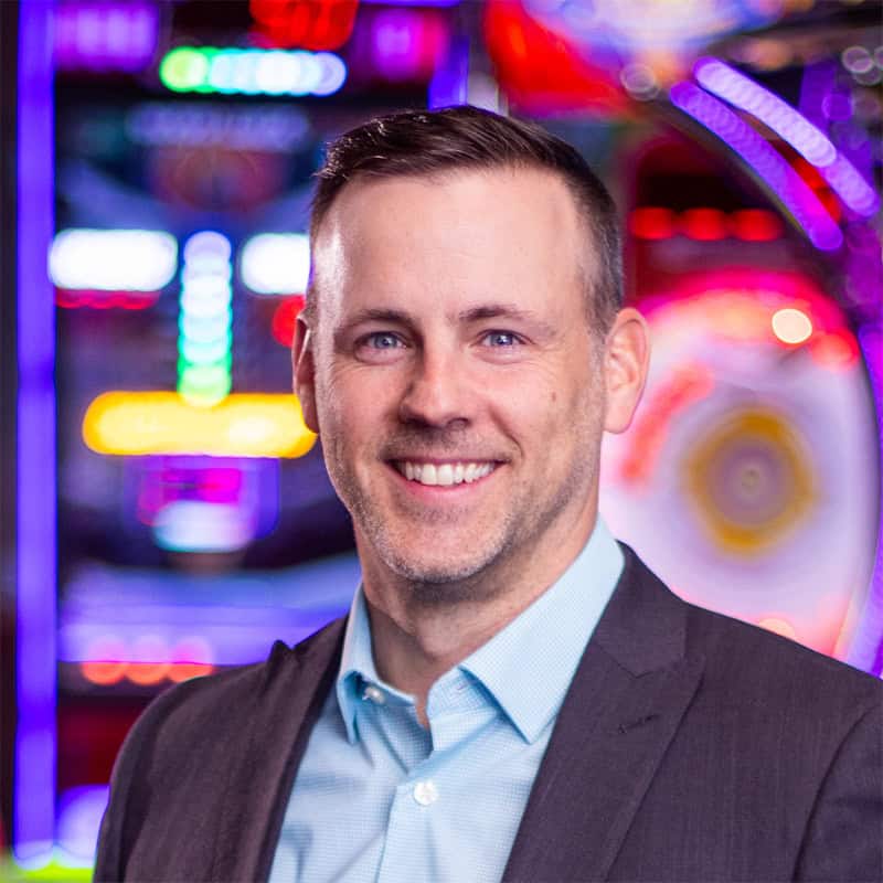 A man in a suit jacket smiles in front of arcade games for kids at Chuck E. Cheese family fun center, known for pizza and birthday parties.
