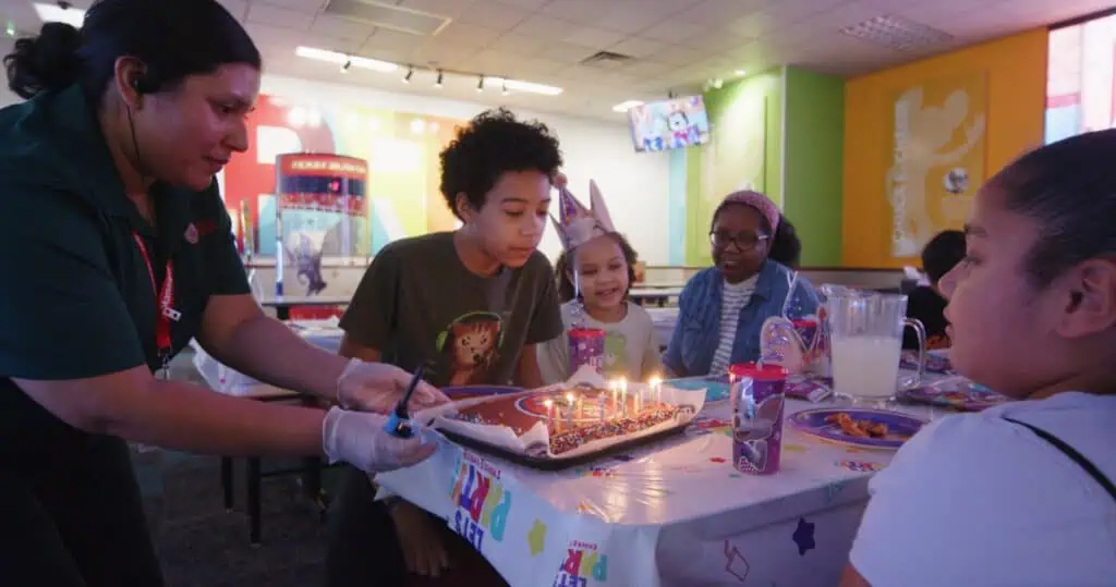 Kids at a Chuck E. Cheese birthday party gather around cake as one child prepares to blow out candles, with arcade games nearby.