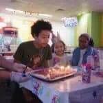 Kids at a Chuck E. Cheese birthday party gather around cake as one child prepares to blow out candles, with arcade games nearby.