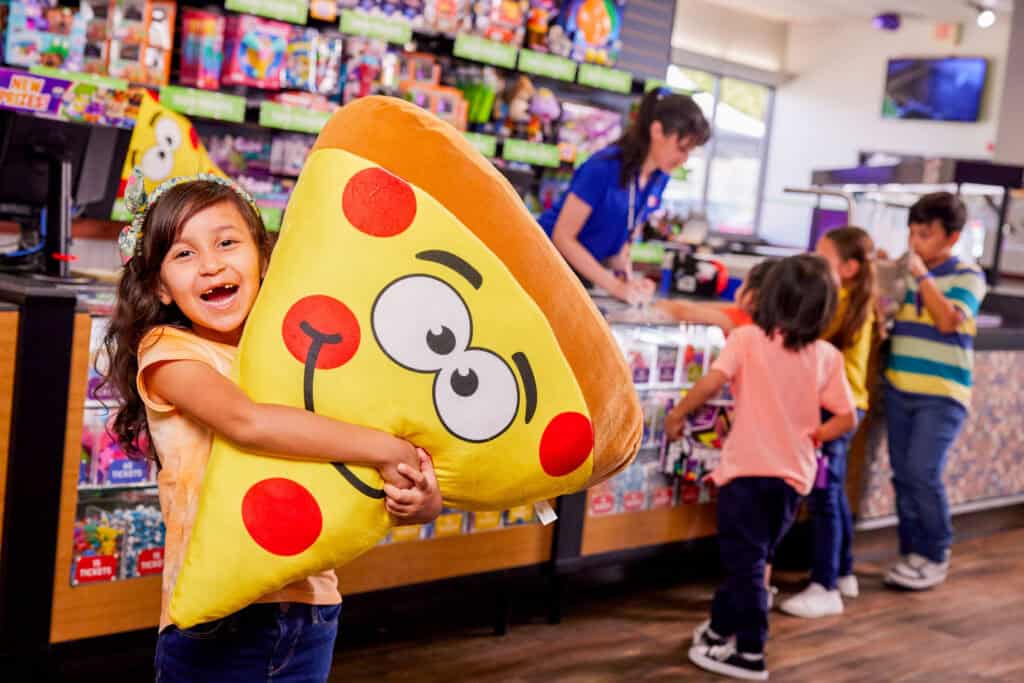 Girl hugs plush pizza slice at Chuck E. Cheese prize counter; other kids and cashier nearby, arcade games in background.