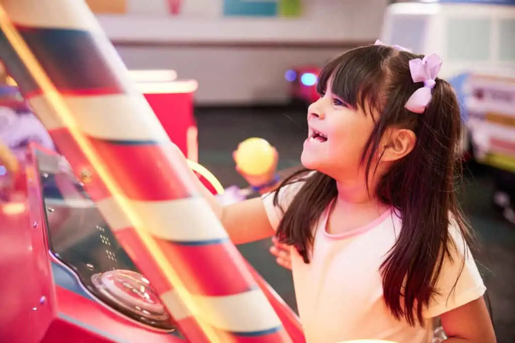 Smiling girl, about 7, plays arcade game at Chuck E. Cheese family fun center, surrounded by colorful lights and game machines.