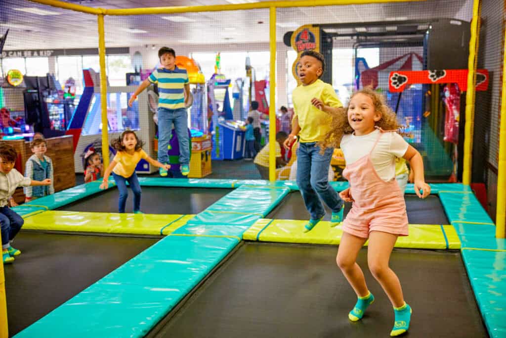 Kids ages 6–10 jump on trampolines in Chuck E. Cheese arcade; others relax by arcade games at the family fun center.