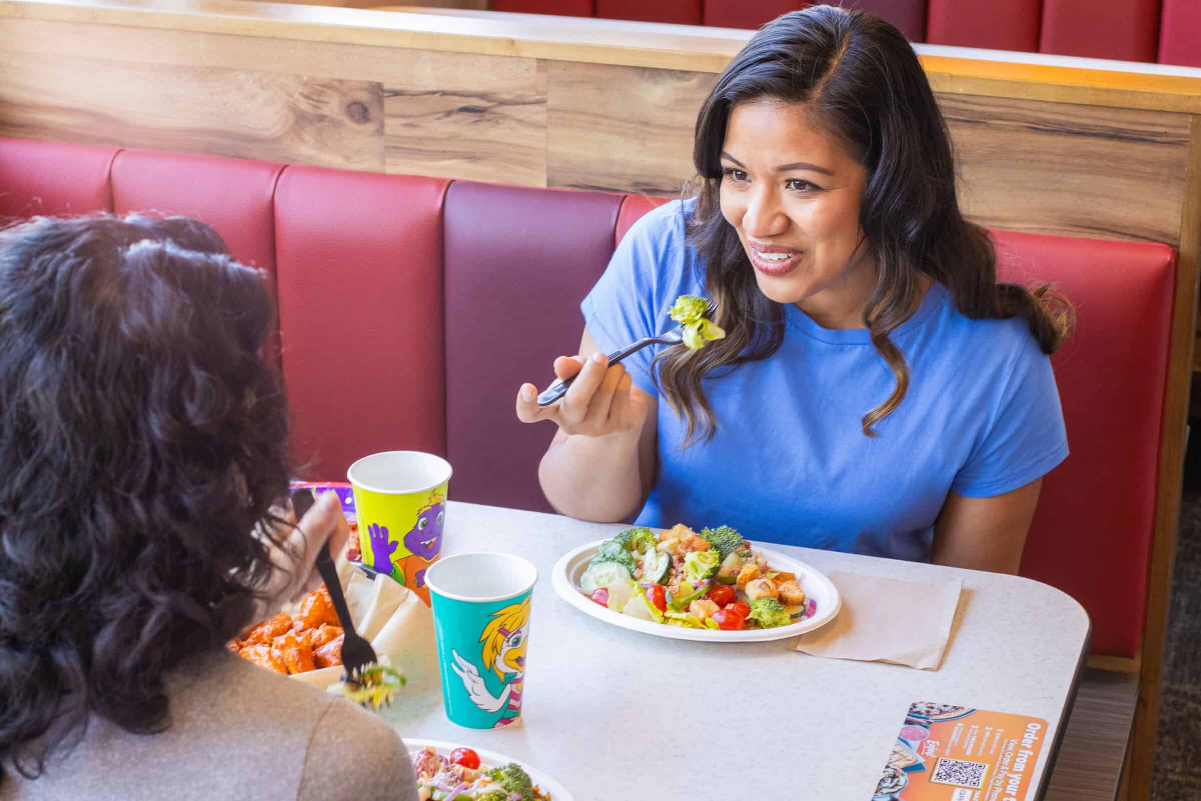 Two women chat and eat salads at a Chuck E. Cheese booth during a kids' birthday party, pizza and drinks on the table.