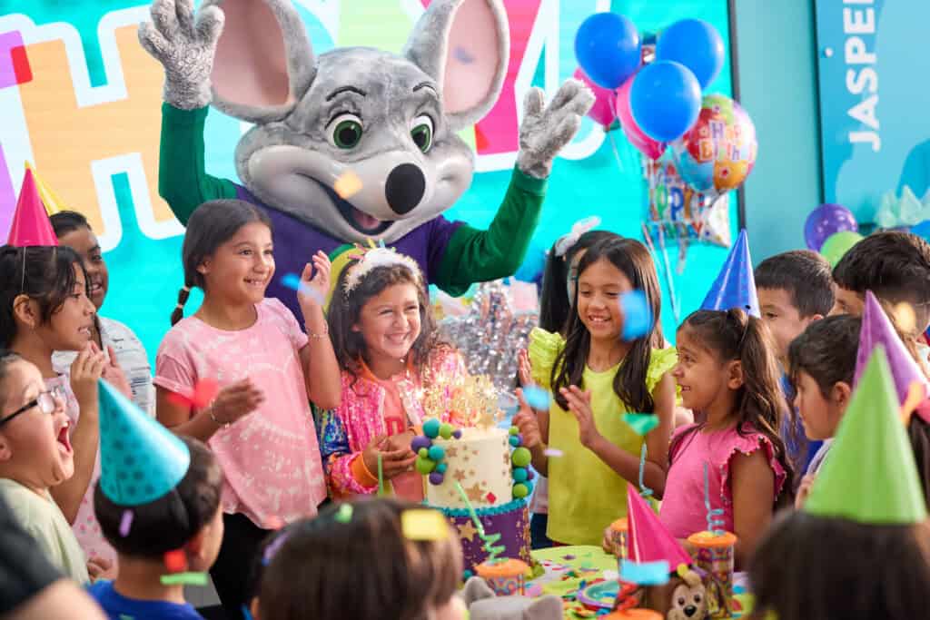 Children celebrating a birthday with Chuck E. Cheese, sparklers on cake, confetti and balloons in the party room.