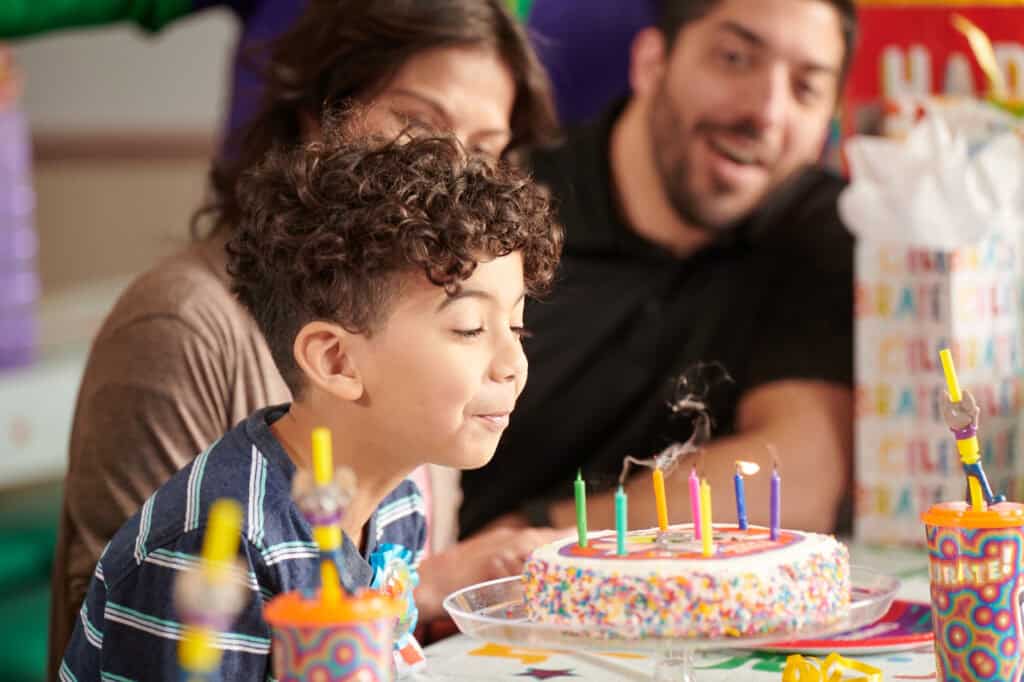 Birthday party in Chuck E. Cheese party room; boy (5–7) blows out candles, adults smile, pizza, arcade games, gifts on table.