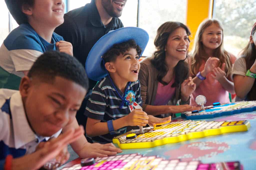 Kids and adults play arcade games in the Chuck&nbsp;E.&nbsp;Cheese arcade area, smiling together at colorful consoles with Fun&nbsp;Pass cards.