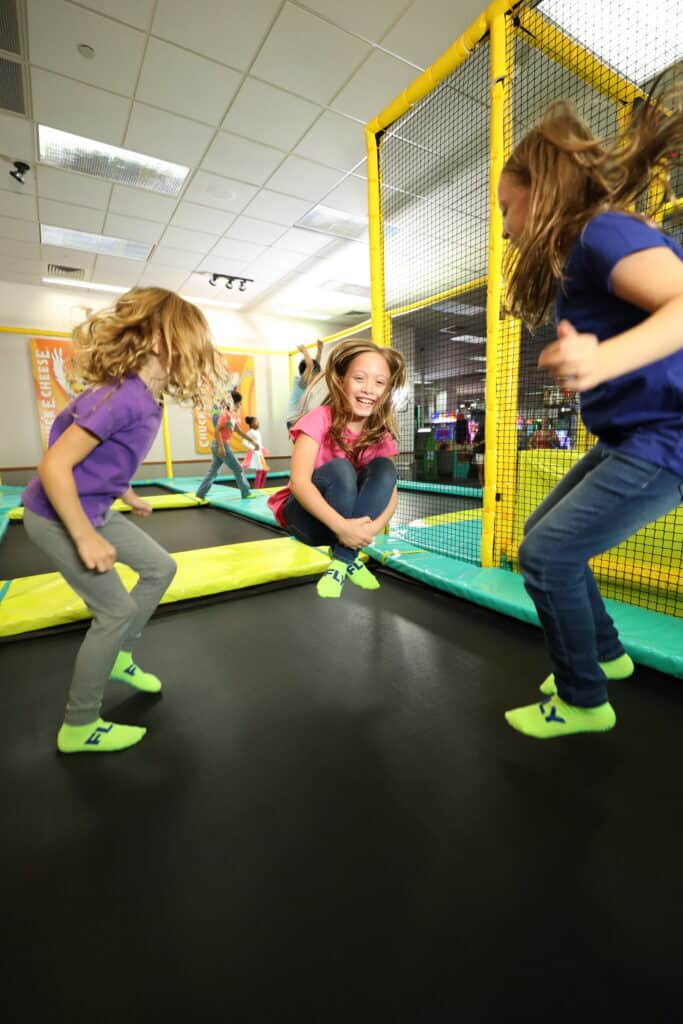 Three girls jump and play on an indoor trampoline at Chuck&nbsp;E.&nbsp;Cheese, surrounded by yellow netting and arcade games
