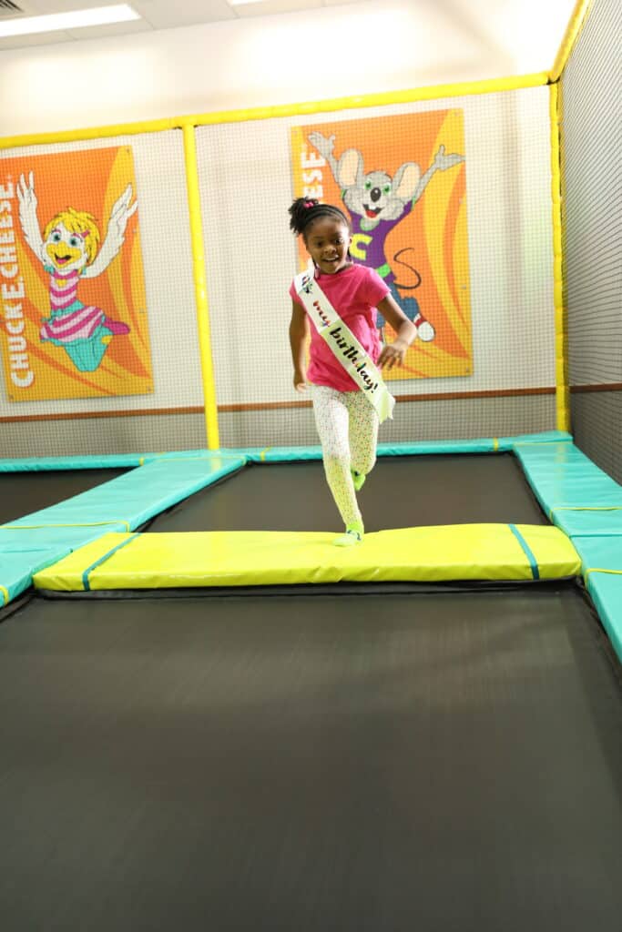 Girl in a birthday sash runs on trampoline at Chuck&nbsp;E.&nbsp;Cheese, with arcade games and safety netting visible