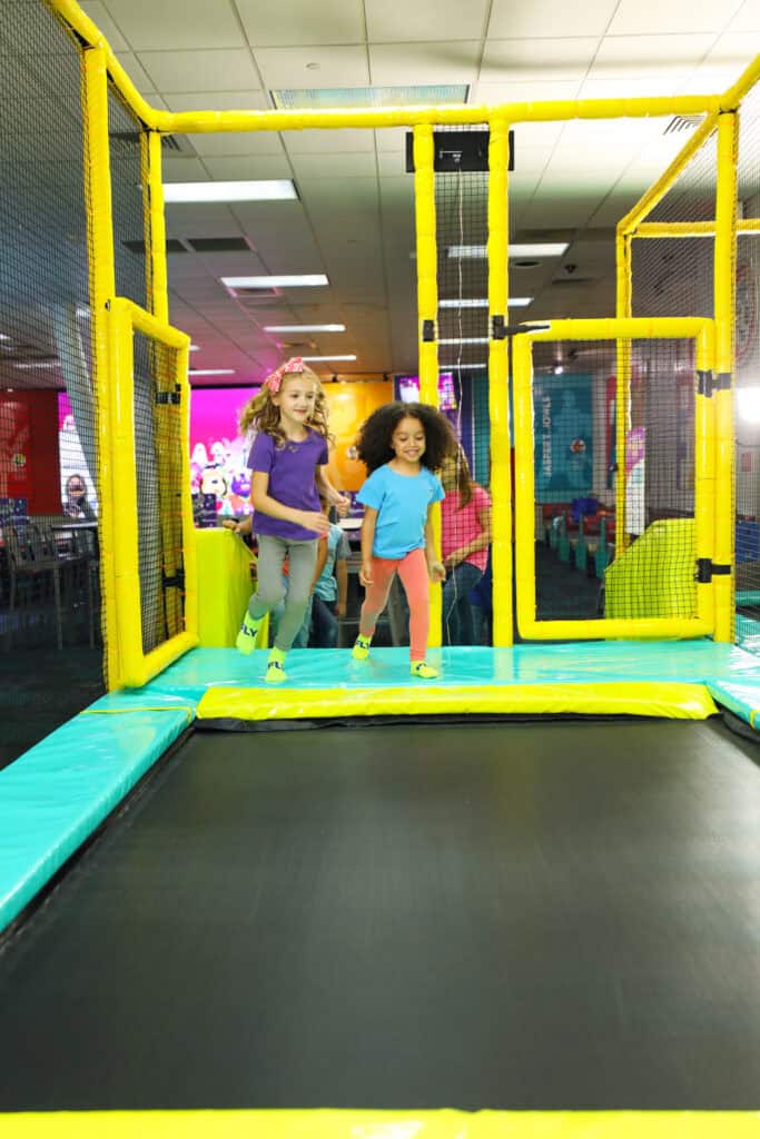 Two girls running on a trampoline at Chuck&nbsp;E.&nbsp;Cheese kids' birthday party, surrounded by arcade games and colorful decor