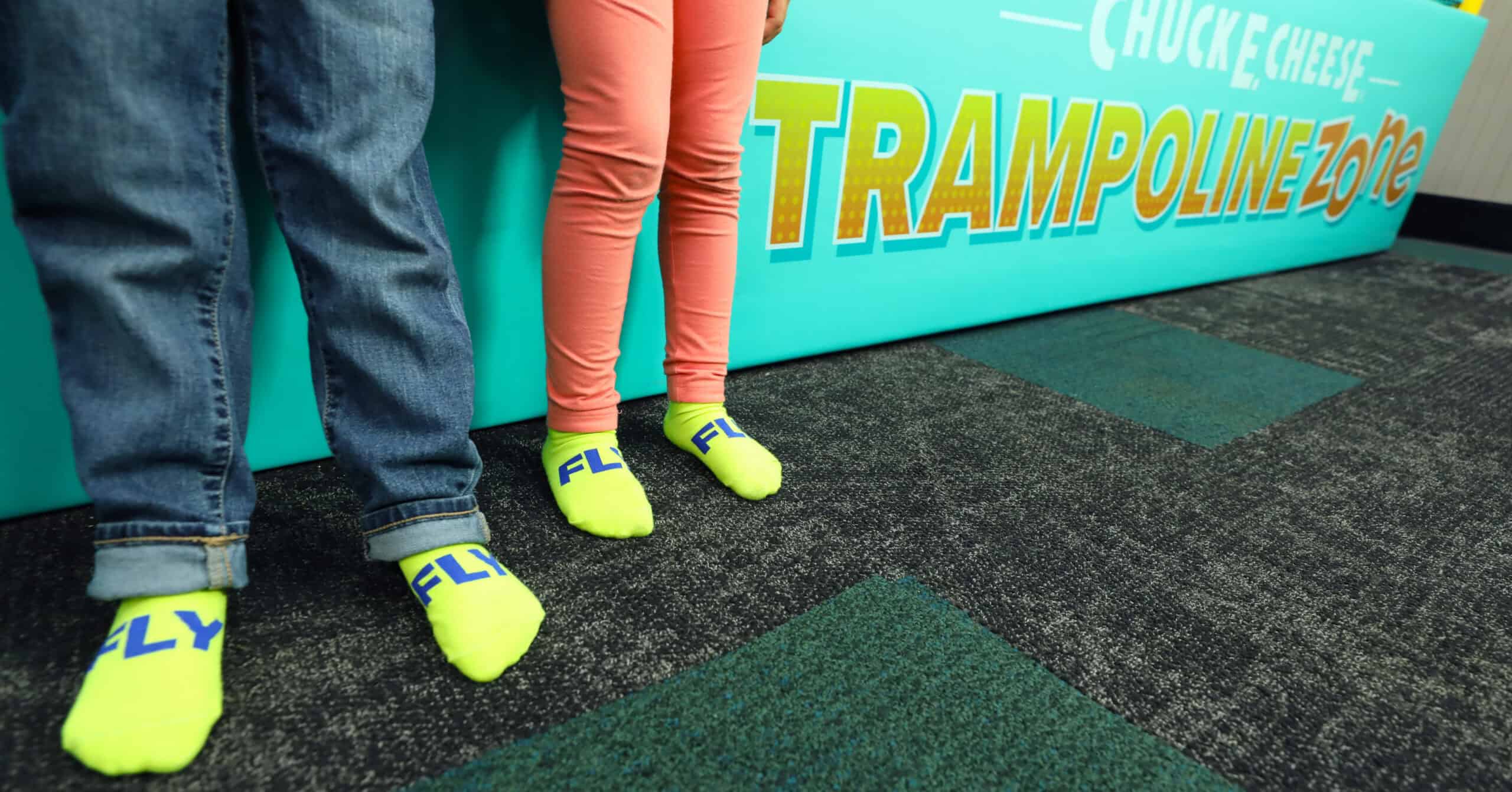 Kids wearing yellow FLY socks stand on carpet by a Chuck&nbsp;E.&nbsp;Cheese Trampoline Zone sign