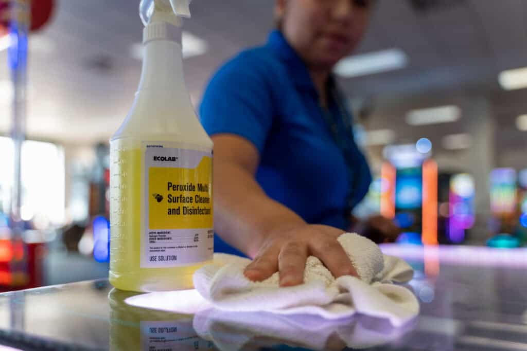 An employee in a blue shirt cleans a Chuck E. Cheese party table with disinfectant; arcade games visible in the background.
