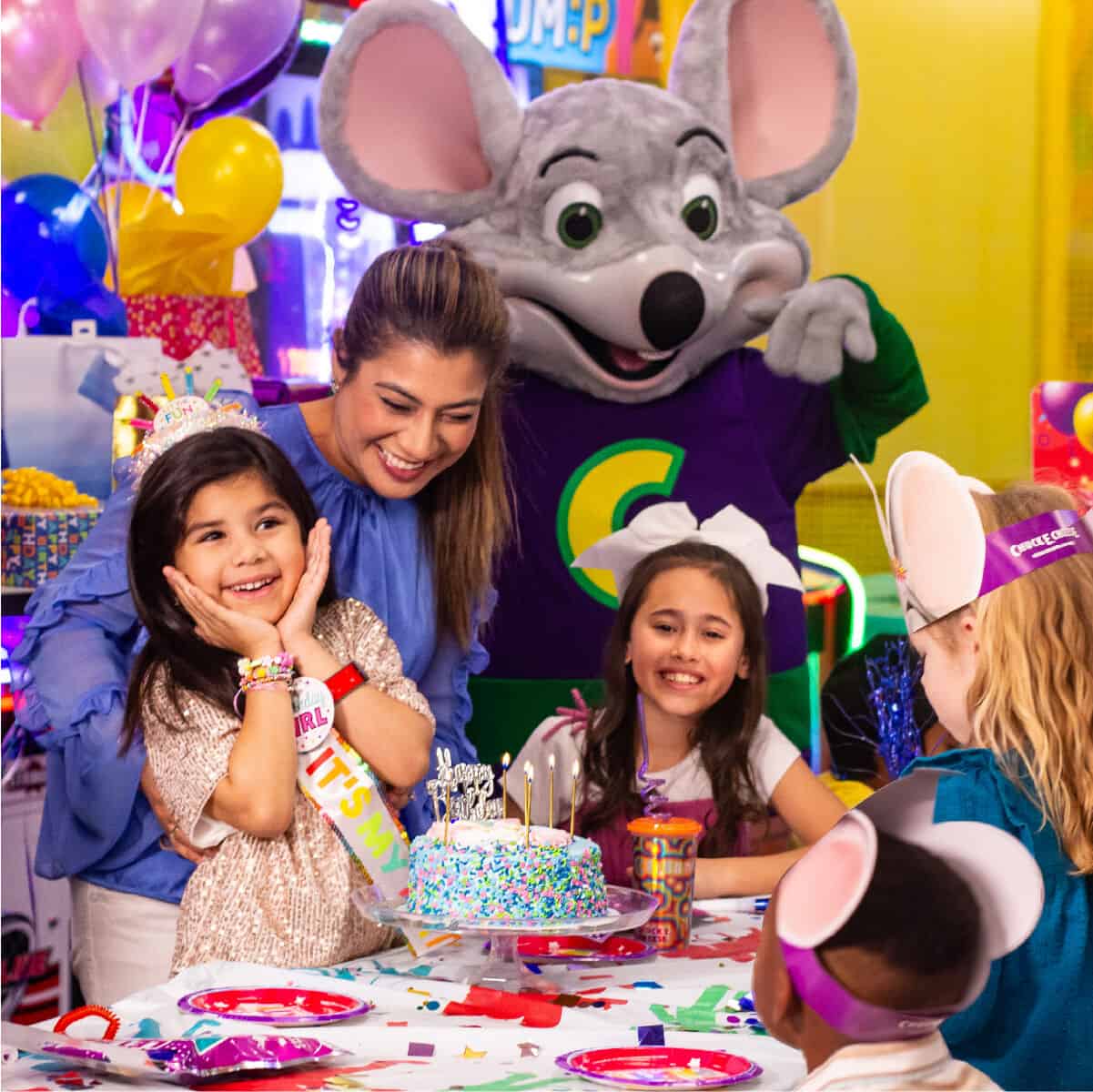 Chuck E. Cheese greets kids ages 5–8 at a birthday party table with cake, balloons, pizza, and arcade games in the party room.