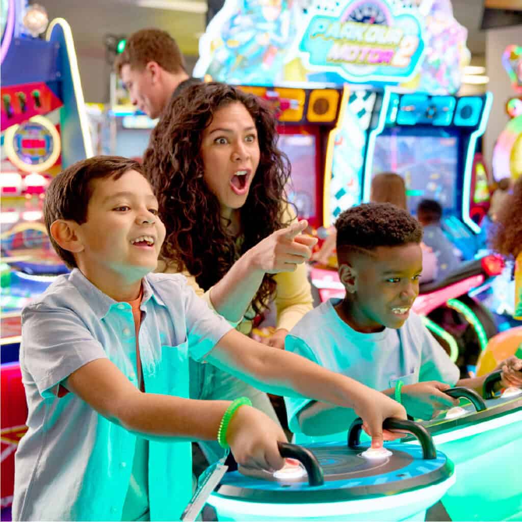 Kids ages 6–9 race arcade driving games at Chuck E. Cheese family fun center arcade, colorful lights, pizza and Fun Pass nearby.
