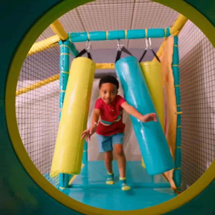 Child in red shirt and blue shorts runs through Chuck E. Cheese play tunnel at family fun center, seen from circular entrance.