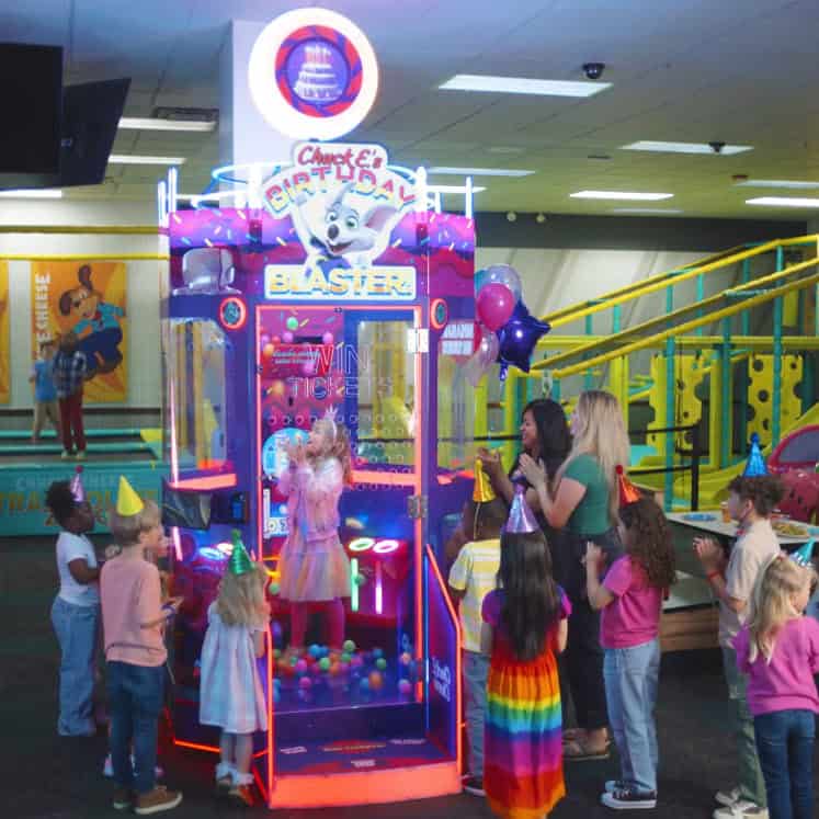 Kids in party hats play the Birthday Blaster arcade game at Chuck E. Cheese birthday party; families watch in a bright fun center.