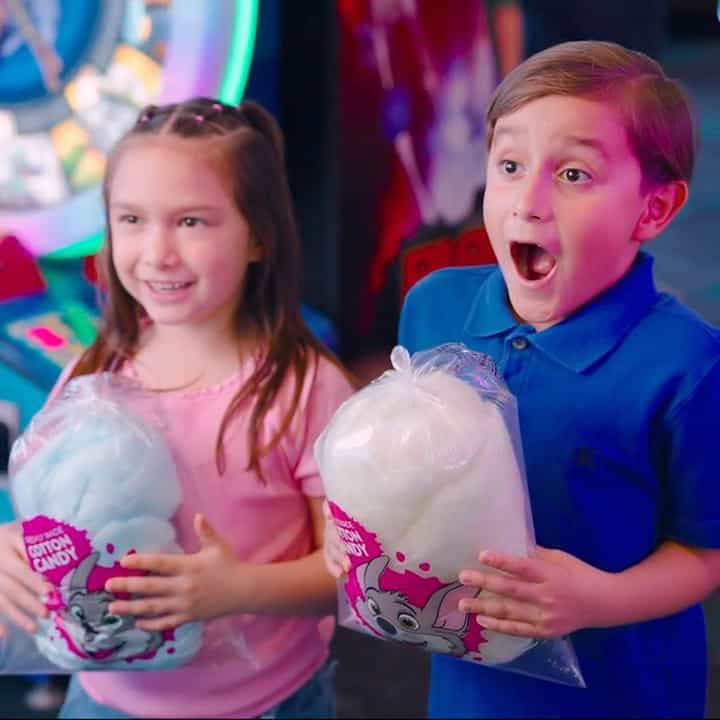 Girl in pink shirt and boy in blue shirt hold cotton candy at Chuck E. Cheese, smiling near arcade games for kids and bright lights.