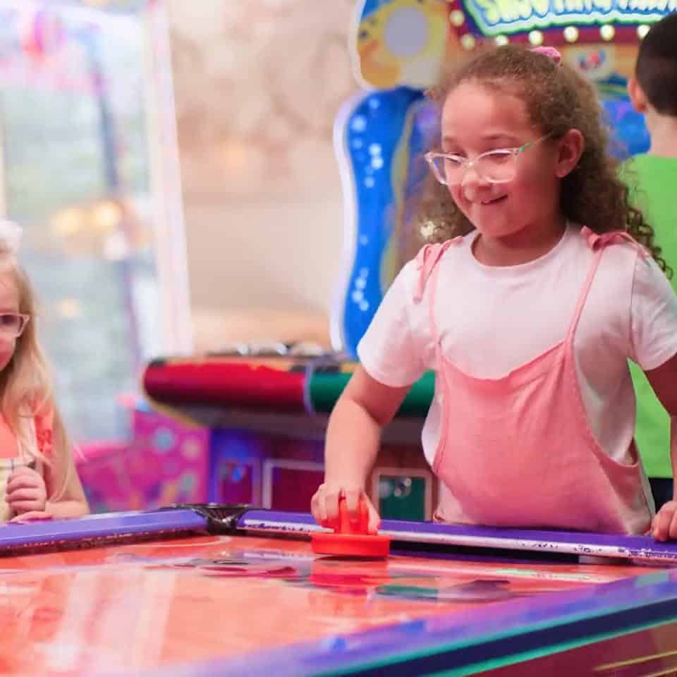 Two young girls play air hockey at Chuck E. Cheese, surrounded by arcade games for kids; one smiles and holds the striker.