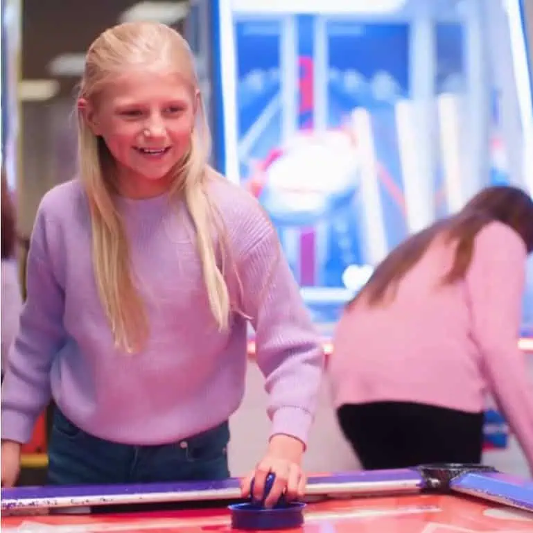 Girl with long blonde hair in lavender sweater plays air hockey at Chuck E. Cheese; another child plays arcade games in back.