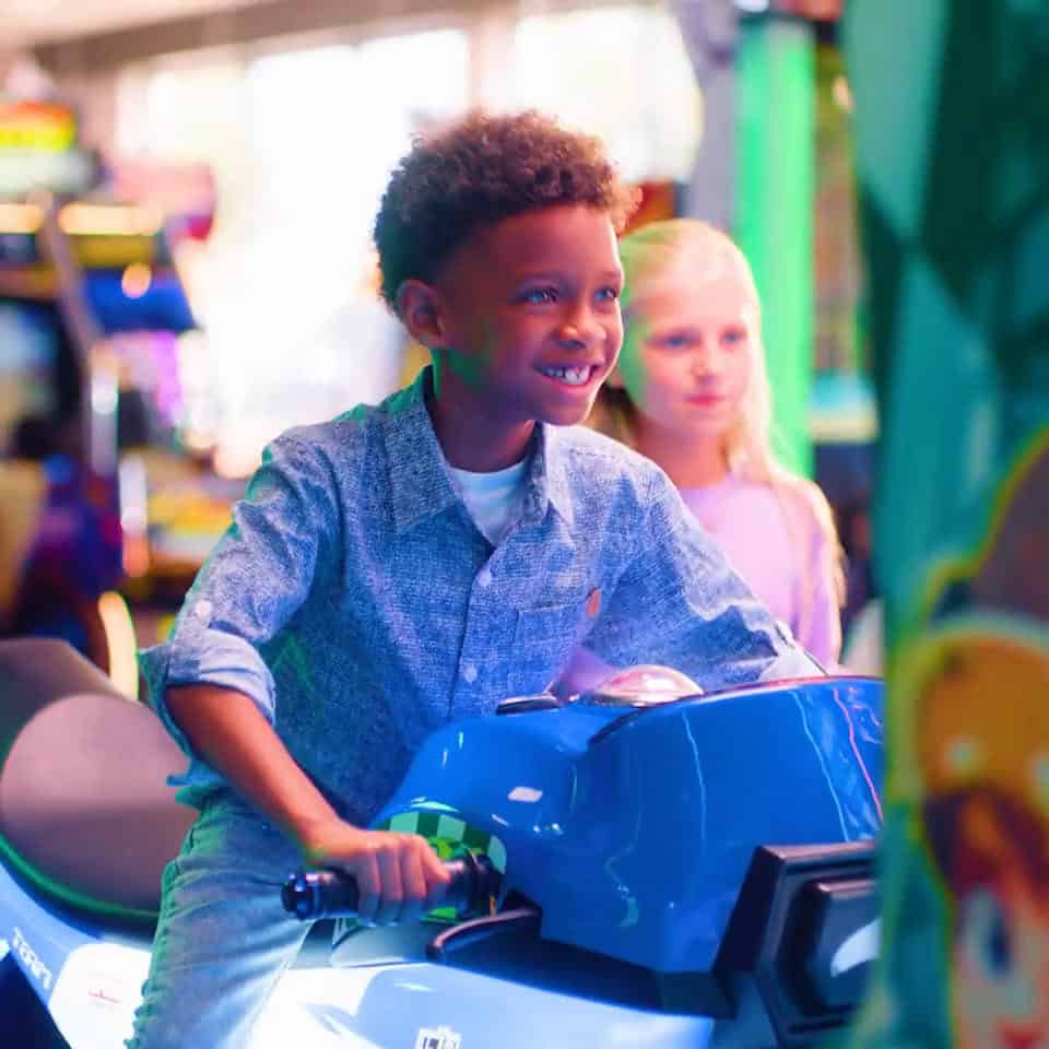 Boy rides a blue motorcycle arcade game at Chuck E. Cheese; girl stands behind, colorful lights, kids birthday party atmosphere.
