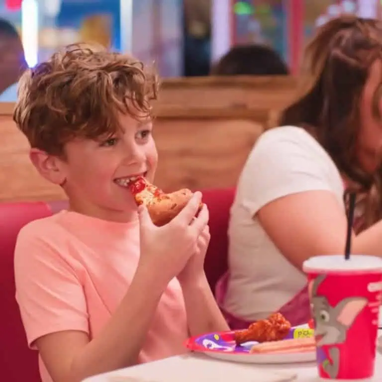 Young boy with curly hair eats pizza at Chuck E. Cheese booth with a woman, red cup, and Fun Pass on table during kids birthday party.