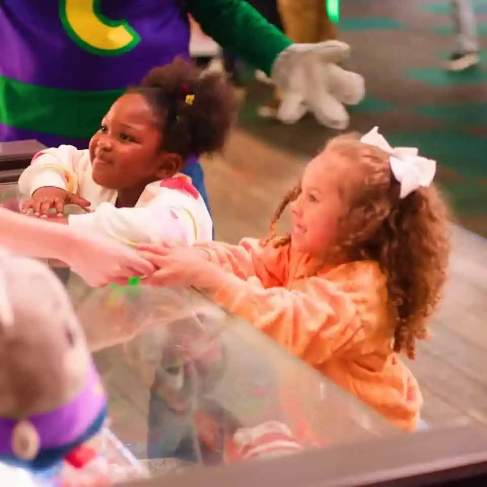 Two young girls at a Chuck E. Cheese birthday party reach for prizes, with Chuck E. in costume behind the counter and games nearby.