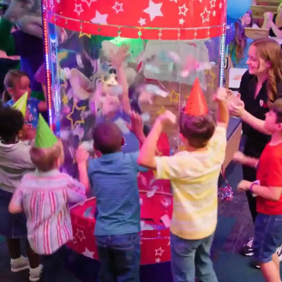Kids in party hats grab swirling tickets in a Chuck E. Cheese booth as an adult supervises a birthday party with pizza and games.