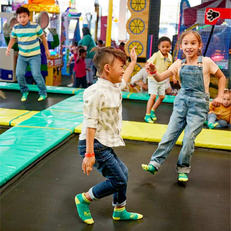 Kids wearing colorful socks jump on trampolines at Chuck E. Cheese, with arcade games and party tables in the background.