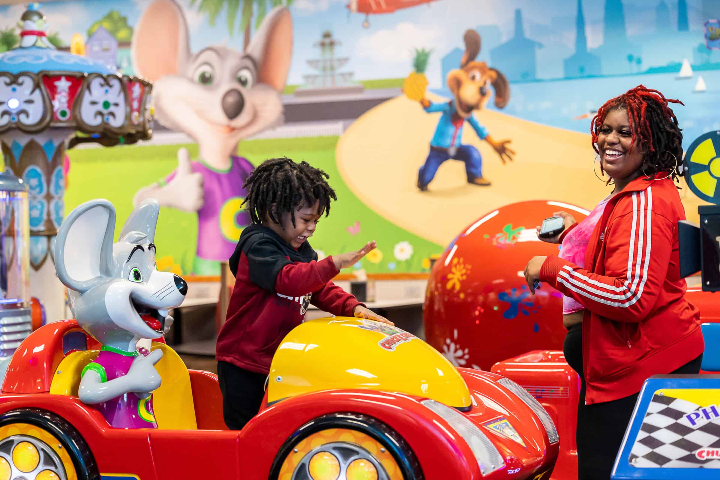 Child playing in a Chuck E. Cheese ride-on game while a woman smiles nearby.